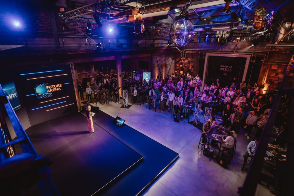 Auf einer Bühne in Future Arena des MuV Forward 25 im Kraftverkehr Chemnitz präsentiert eine Moderatorin dem Publikum das Programm